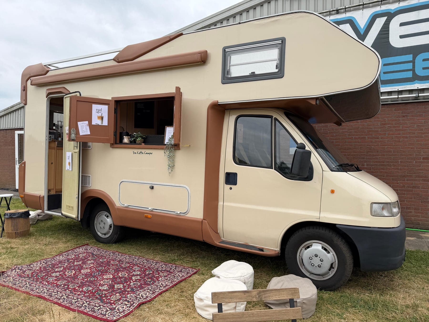 De Koffie Camper met open luik, kleed en poefs op het gras
